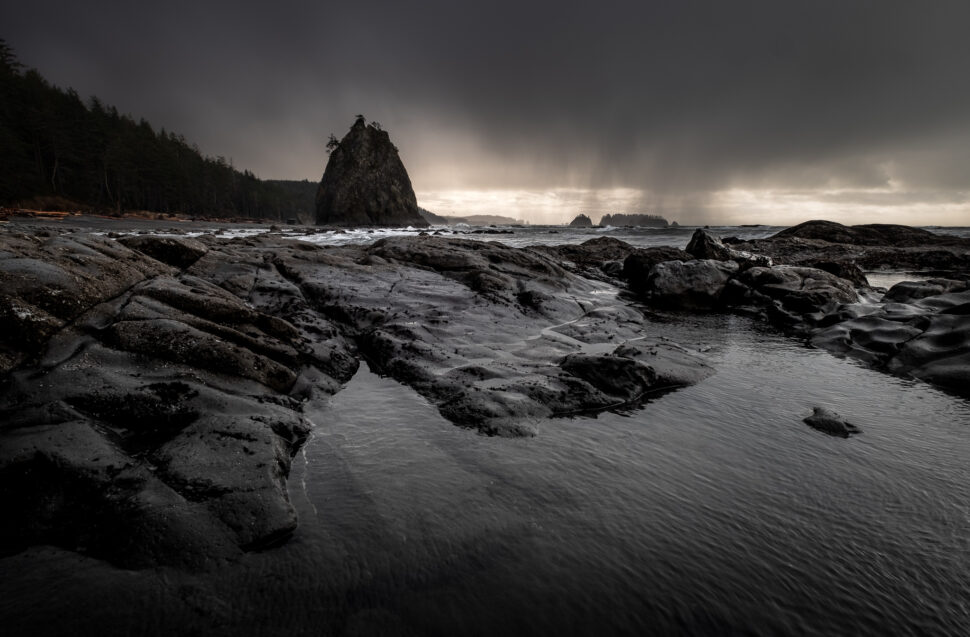 Dark, jagged rocks along a stormy shoreline with a large sea stack and misty rain in the distance; rough water pools reflect the overcast sky.