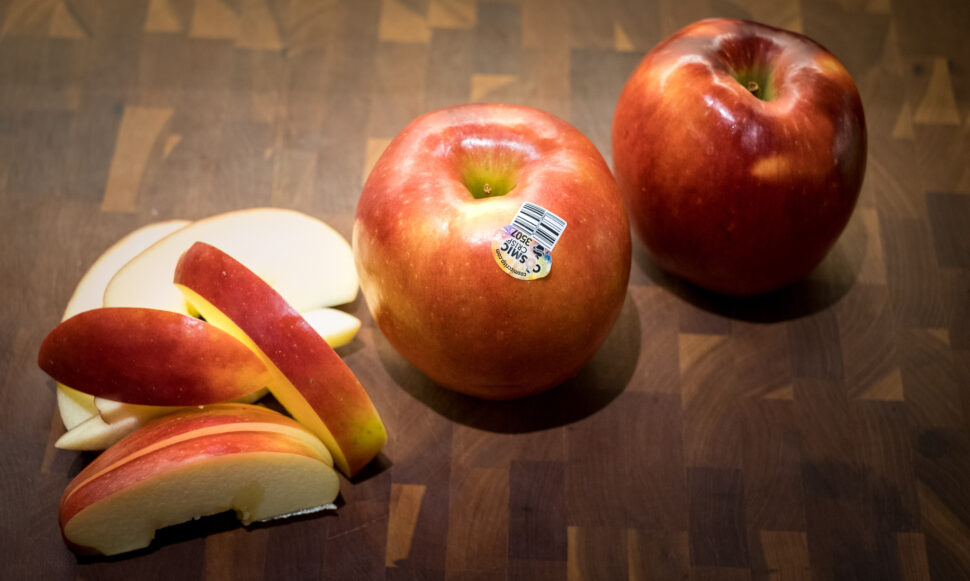 Two whole red apples, one with a sticker, sit on a wooden cutting board next to a group of sliced apple pieces in the foreground.