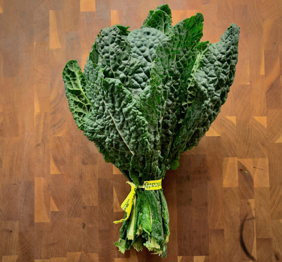 Bundle of dark green kale with crinkled leaves tied with a yellow band, placed on a wooden floor.